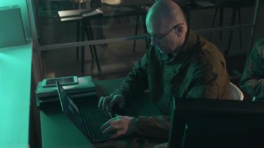 Beyaz erkek askeri gözetim memurunun yüksek açılı görüntüsü. Masada oturmuş dizüstü bilgisayarına bir şey yazarken ve gece bilgisayar ekranına bakarken.