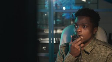 Orta ölçekli Afrika kökenli Amerikan askeri gözetim subayı bilgisayar ekranlarının önünde oturmuş onlara bakıyor ve telsizle rapor gönderiyor.