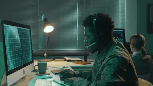 非洲裔美国男性军事监视官坐在办公桌前 用雷达盯着电脑屏幕 用耳机交谈 并做笔记 — 图库视频影像