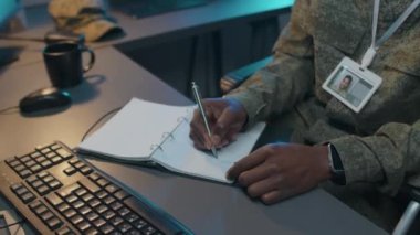 Odaklanmış erkek Afro-Amerikan ordu subayının fotoğraf çekimi. Masa başında oturmuş, deftere not alıyor, bilgisayar klavyesine yazı yazıyor ve kulaklıkla konuşuyor.
