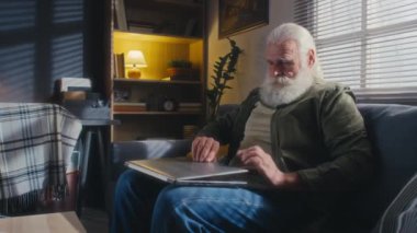 Beyaz saçlı, kalın sakallı ve bıyıklı bir erkek emeklinin evde tek başına oturup dizüstü bilgisayarını alıp video çağrısına cevap verirken ve biriyle konuşurken orta ölçekli bir fotoğrafı.