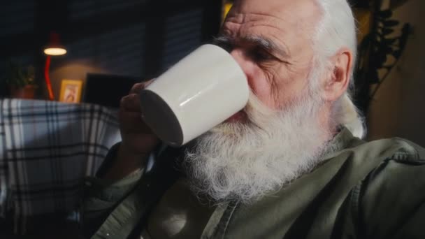 一位体贴入微的老人 留着浓密的灰胡子和胡子 在家里喝咖啡 从窗户的百叶窗往外看 拍下了他的近照 — 图库视频影像