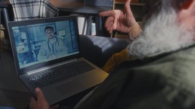 Gri saçlı, sakallı, elinde dizüstü bilgisayarla oturan ve video ekranında pratisyen hekimiyle konuşan tanınmayan bir adamın orta boy yakın çekimi.
