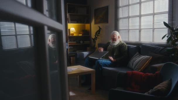 满满一帧退休的白发男子坐在家里的沙发上 拿起笔记本电脑 打开电脑 与医生通过视频通话 描述他的病情和抱怨 — 图库视频影像