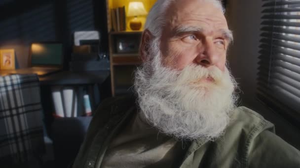 留恋的老年男性鳏夫 灰白的头发 浓密的胡须和胡子 坐在窗前 凝视着外面 然后是相机和微笑的特写镜头 — 图库视频影像