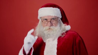 Noel Baba 'nın kırmızı ceketli, siyah şapkalı ve gözlüklü, gri saçlı, sakallı ve bıyıklı, kameraya bakan ve kötü davranışa ve itaatsizliğe parmak sallayan orta boy yakın çekimi.