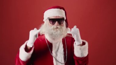 Hipster Noel Baba 'nın kırmızı kostümlü ve güneş gözlüklü, boynunda gümüş zincir olan ve boynuzlu el hareketleriyle dans eden orta boy yakın çekimi. Şablon, boşluğu kopyala