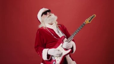 Noel Baba kıyafetli, güneş gözlüklü, gümüş zincirli, elektro gitar çalan, sallanan ve şarkı söyleyen orta yaşlı bir adamın resmi. Şablon kadar uygun, alanı kopyala