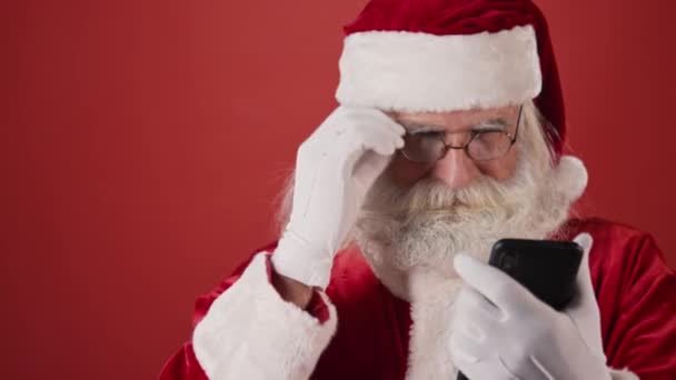 中等比例的大胡子老人身穿圣诞老人服装 看着智能手机 摘下眼镜 眯着眼 阅读短信 然后带着喜悦和喜悦微笑 复制空间 — 图库视频影像