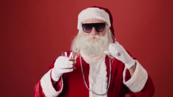 穿着圣诞老人外套 戴着太阳镜 戴着银链 留着胡子 喝着威士忌 跳着时髦的鸡舞的中杯老年男子 背景是浅红的 复制空间 — 图库视频影像