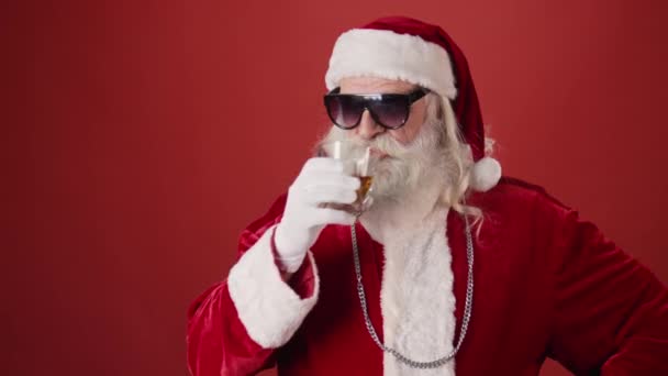 身着圣诞老人服装 头戴帽子 头戴银链子 留着胡子和胡子的中档老人的照片 他们喝着啤酒瓶里的威士忌 摘下太阳镜 眨着眼睛 红色背景 复制空间 — 图库视频影像