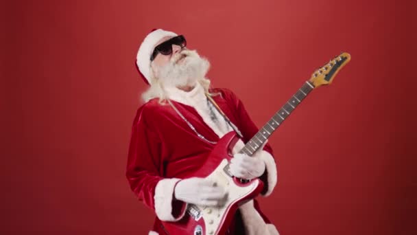身着圣诞老人服装 头戴墨镜 戴着银链 弹奏电吉他 摇动着 背景为红色 适用于模板 复制空间 — 图库视频影像