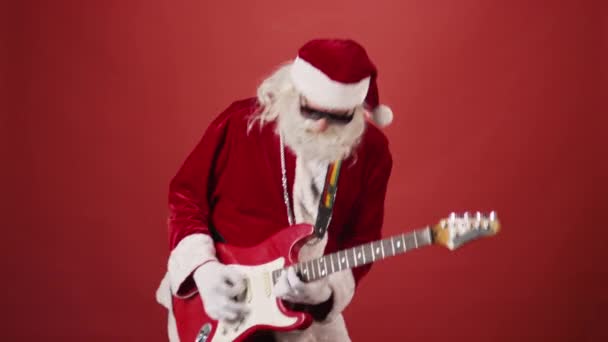 中镜头圣诞老人身穿红色服装 太阳镜和银链 弹奏电吉他 伸出舌头 红色背景 复制空间 — 图库视频影像