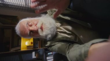 Gülümseyen, gri saçlı, sakallı ve bıyıklı yaşlı bir adamın orta boy dikey görüntüsü. Kanepede oturup torunları ya da ailesi için akıllı telefondan selfie videosu kaydediyor.