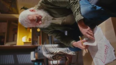Orta boy, gri saçlı, beyaz sakallı ve bıyıklı yaşlı bir adamın küçük bir dairede tek başına oturup ödenmemiş ve gecikmiş faturaları incelerken çekilmiş dikey bir fotoğrafı.