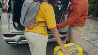 Kafkasyalı genç kadın ve Hintli kocanın caddede durup büyük bavulları arabanın bagajına doldurup tatile çıkmaya hazırlanırken orta yavaş çekimleri.