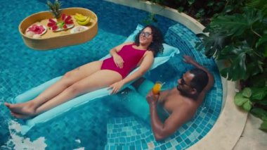 Beyaz bir kadının yüzme havuzunda yüzerken, afro-amerikalı bir adamla sohbet ederken, yaz tatilinde lüks egzotik bir otelde dinlenirken tam açılı bir pozu.