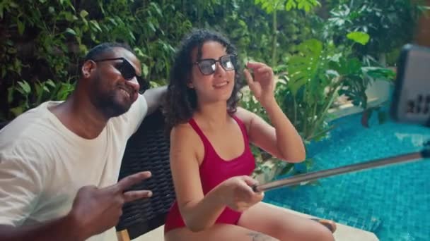 在国外度假期间 一对多民族夫妇坐在豪华酒店的游泳池边 手持智能手机自拍 看着相机 并以滑稽的姿势笑着 — 图库视频影像