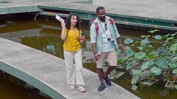 アフリカ系アメリカ人男性と白人女性のフルショット バックパックと観光パンフレットは 歴史的なランドマークを訪問した後 公共公園の池の上の徒歩パスで一緒に歩きます 笑顔とチャット — ストック動画