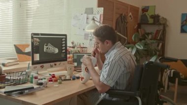 Orta boy beyaz erkek beyaz tasarımcı tekerlekli sandalyede bilgisayarın önünde oturuyor ve gündüz stüdyoda beyaz ayakkabıları özelleştiriyor.