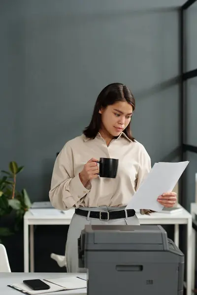 Young brunette woman in quiet luxury attire looking through financial document and having cup of coffee in office while standing by copier
