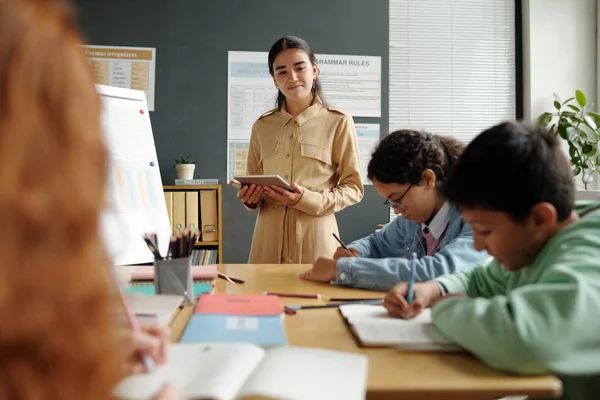 タブレットを使用し 新しい文法規則でホワイトボードで立っている間 コピーブックでノートを作る生徒のグループを見る若い教師 — ストック写真