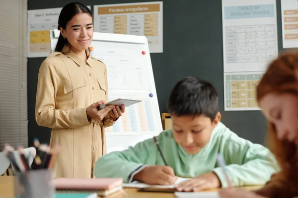 Young Smiling Teacher Tablet Looking One Several Diligent Schoolkids Writing — Stock Photo, Image