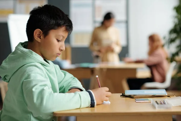 Side view of serious multiethnic schoolboy in casualwear making notes in copybook while sitting in front of camera by desk at lesson of English