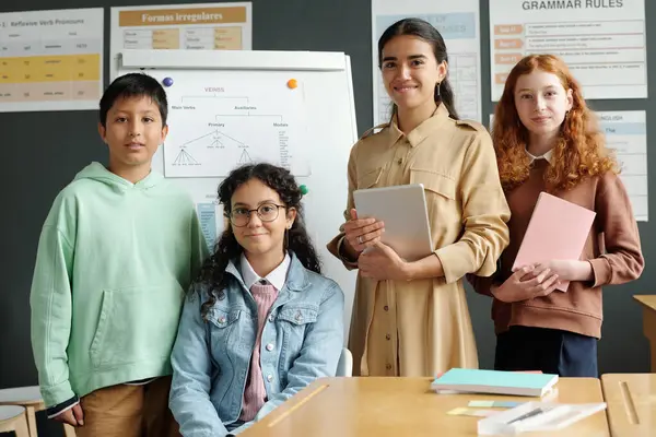 タブレット付きのハッピーな若い英語教師とデスクで立ってカメラを見ている中学校の異文化学習者のグループ — ストック写真