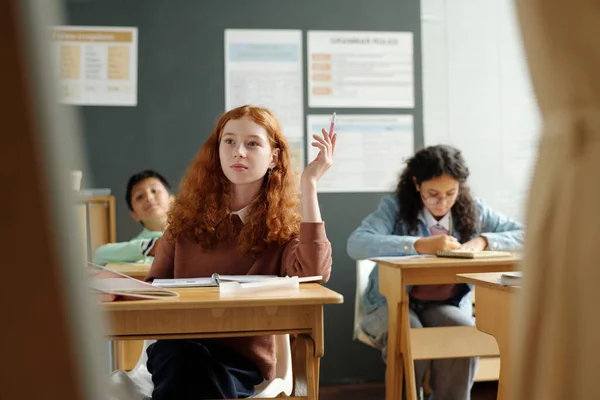 卓越した女子高生が机に座りながら手を挙げ 英語文法の新しいルールについて議論する際にホワイトボードを見る — ストック写真