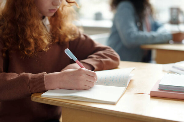 Hand of diligent schoolgirl with pen writing down new grammar rules of English language while sitting by desk at lesson against classmate