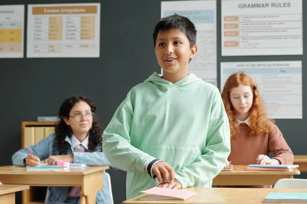 Happy schoolboy in hoodie standing by desk and answering question of teacher at lesson of English grammar against two schoolgirls