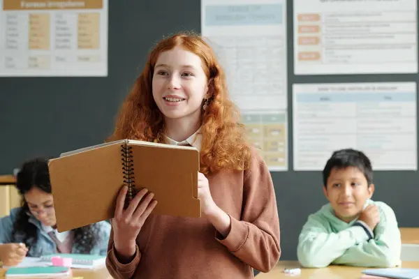 Clever schoolgirl with long ginger wavy hair answering question of teacher at lesson of English language while holding open copybook with notes