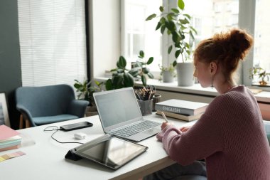 Öğrenci kız notlar alırken ve öğretmenlerin yeni kuralları açıklarken online ders sırasında dizüstü bilgisayarın önünde otururken yan görünümü