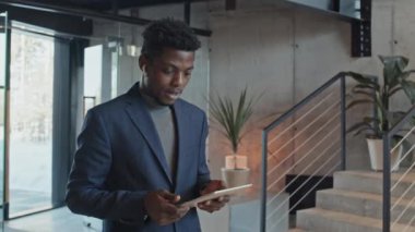 Orta boy Afro-Amerikan erkek şirket çalışanı ofis lobisinde kablosuz kulaklıkla konuşuyor ve iş toplantısına giderken tablet bilgisayara bakıyor.