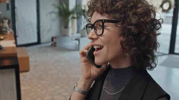 カーリーヘアの若い白人女性の顔のクローズアップアークスローモーショット ホテルやオフィスロビーでスマートフォンで話すメガネでポジティブな感情 — ストック動画