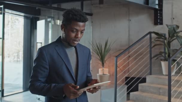 オフィスロビーに立ってスーツを着た若いアフリカ系アメリカ人男性企業の従業員の中型ショット ワイヤレスイヤホンを通して話し ビジネスミーティングに向かう途中のタブレットコンピュータを見る — ストック動画