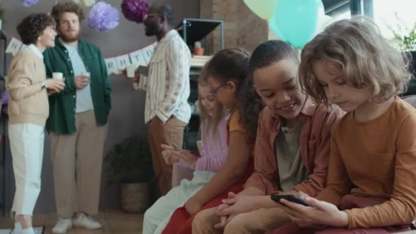 若い白人と黒人の少年少女が誕生日のパーティーでソファーに座り スマートフォンを見たり ブラウジングやチャットしたり 両親がアニメーションの会話をしている最中 — ストック動画