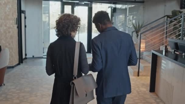 後部は オフィスドアに向かって一緒に歩いて タブレットコンピュータを見て ビジネスミーティングへの途中でデータを議論するスーツにカーリーコーカサス女性と黒人の男性のショットに従ってください — ストック動画