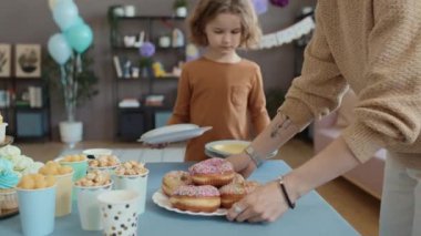 Evde doğum günü partisine hazırlanan, çörekleri, çörekleri, patlamış mısırları masaya koyup servis tabakları hazırlayan tanınmamış kadın ve küçük çocuğun orta yavaş çekimi.