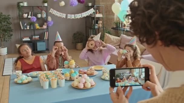 一个不知名的女人手牵着智能手机 拍了许多十几岁以下儿童的照片 在生日派对上端着糖果坐在桌旁 做傻脸 鬼鬼祟祟 — 图库视频影像