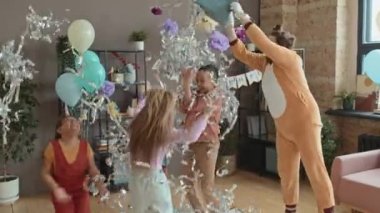 Kedi kostümü giymiş beyaz bir adam doğum günü partisinde çocuklara süs eşyası fırlatıyor ve heyecanlı çocuklar gülüyor, dans ediyor ve onunla oynuyor.