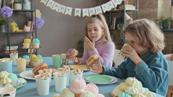在生日宴会上 9岁的白人女孩和男孩一起坐在餐桌前 吃着甜甜圈 爆米花 棉花糖和纸杯蛋糕 聊着天 — 图库视频影像
