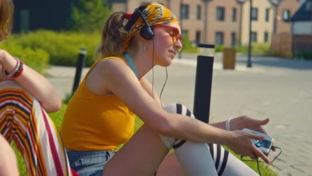 明るいヒッピースタイルのヘッドフォンを着て 友人と緑の芝生に座っている間 古いCdプレーヤーに耳を傾けている若い白人少女のミディアムショット — ストック動画