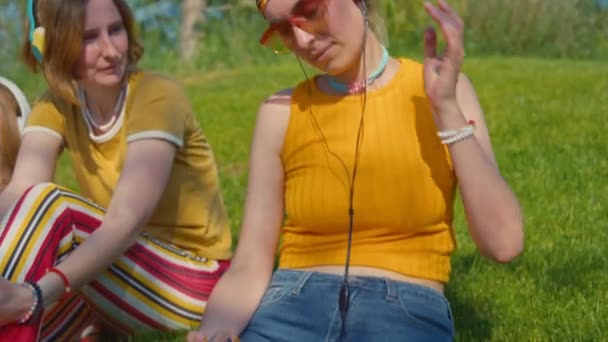 明るいヒッピースタイルのヘッドフォンを着て 友人と緑の芝生に座っている間 古いCdプレーヤーを聴く 明るいヒッピースタイルの若い白人女性のティルトショット — ストック動画