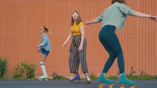 バックグラウンドで屋外クワッドローラーで専門的にスケートするレトロカラフルな服の3人の女の子の安定したフルショット オレンジの壁 — ストック動画