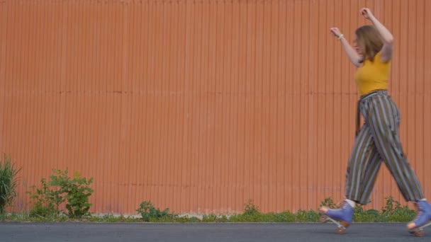レトロな服とクワッドスケートを着た3人の明るい十代の少女の安定したフルショットは 昼間外のオレンジ色の壁の前で次々とポーズし 乗っています — ストック動画