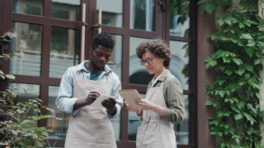 İki çok ırklı kafe çalışanının dışarıda durup notlar alırken ve gündüz çalışma sürecini tartışırken orta boy görüntüsü.
