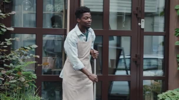 アフリカ系アメリカ人のウェイターがテラスを掃除し 昼間外で誰かに挨拶する中型ショット — ストック動画