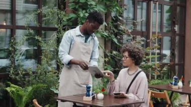 Erkek Afro-Amerikan kafe çalışanı elinde tabletle menü tutarken ve beyaz kadın müşteriye gündüz terasta sipariş verirken yardım ederken orta boy bir fotoğraf.